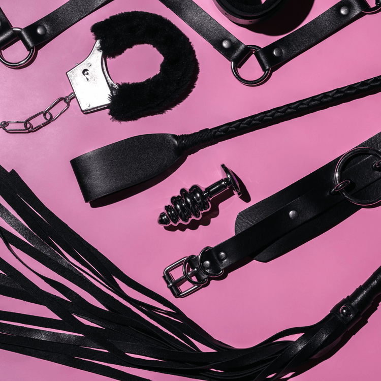 Leather bondage accessories arranged on a pink background, including cuffs, a whip, and a butt plug.