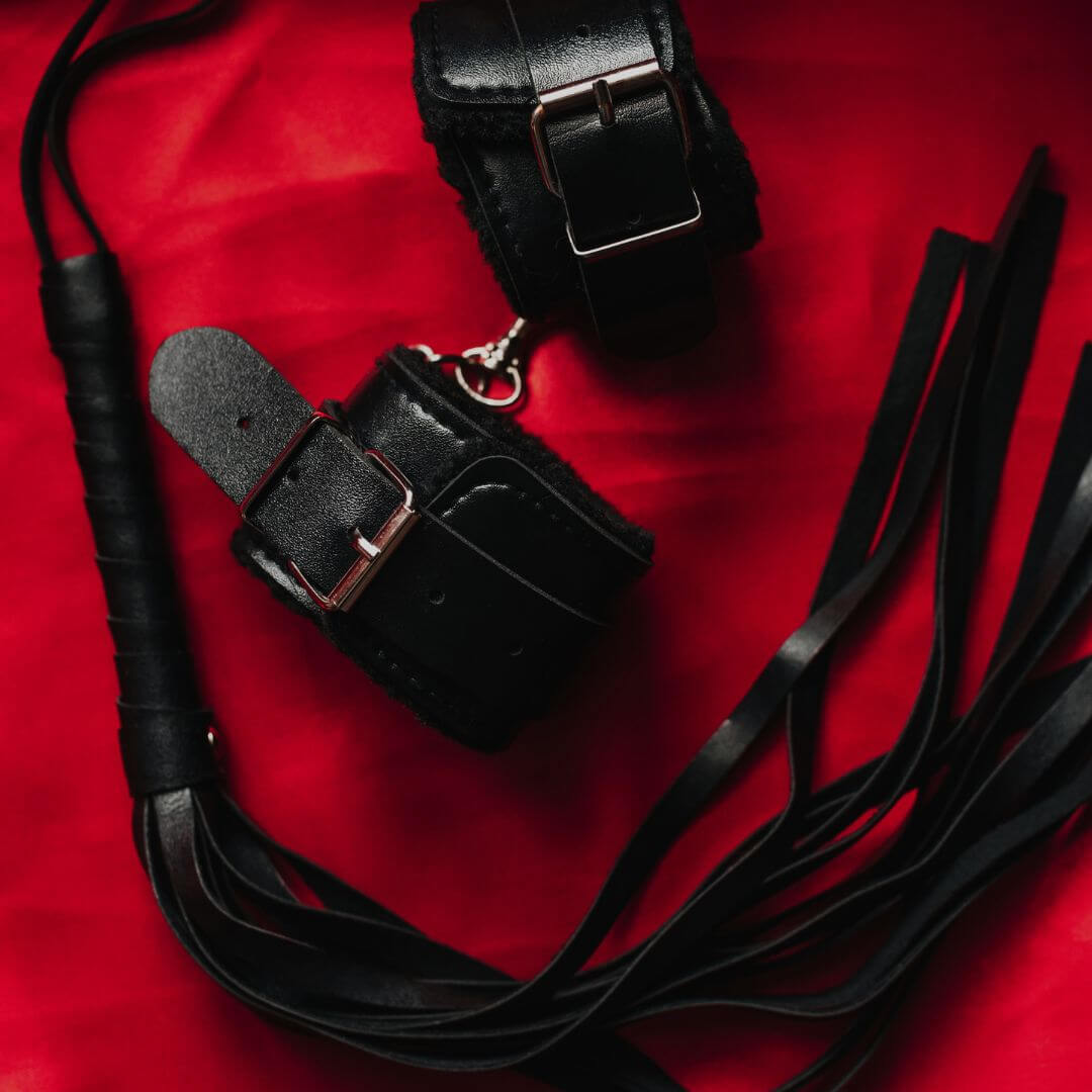 Black leather cuffs and whip on red background, perfect for bondage and BDSM play.