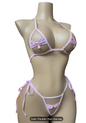 A pink string bikini set displayed on a beige mannequin torso, featuring a triangle bra top with a small bow detail and a matching string thong with side ties and a bow. A Christmas tree and candles are visible on the left against a dark gray background.
